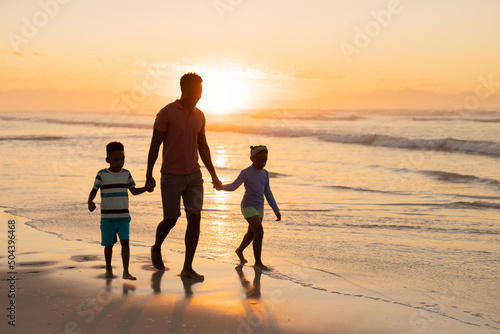 African american young father holding son and daughter's hands while walking on beach against sky