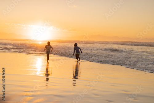 Playful african american brother and sister running at beach against sky during sunset