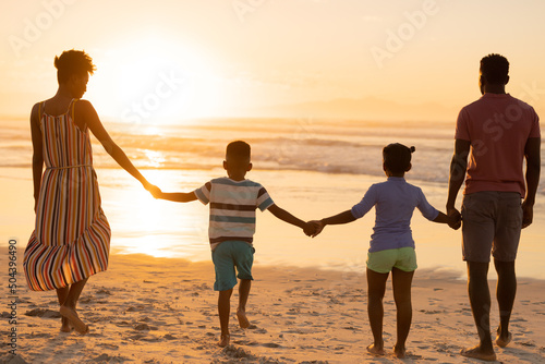 Rear view of african american young parents holding daughter and son's hands while walking on beach