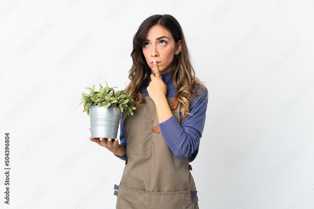 Gardener girl holding a plant isolated on white background having doubts while looking up Gardener girl holding a plant isolated on white background having doubts while looking up