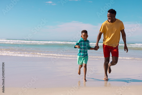 Cheerful african american young man and son holding hands while running at beach against blue sky