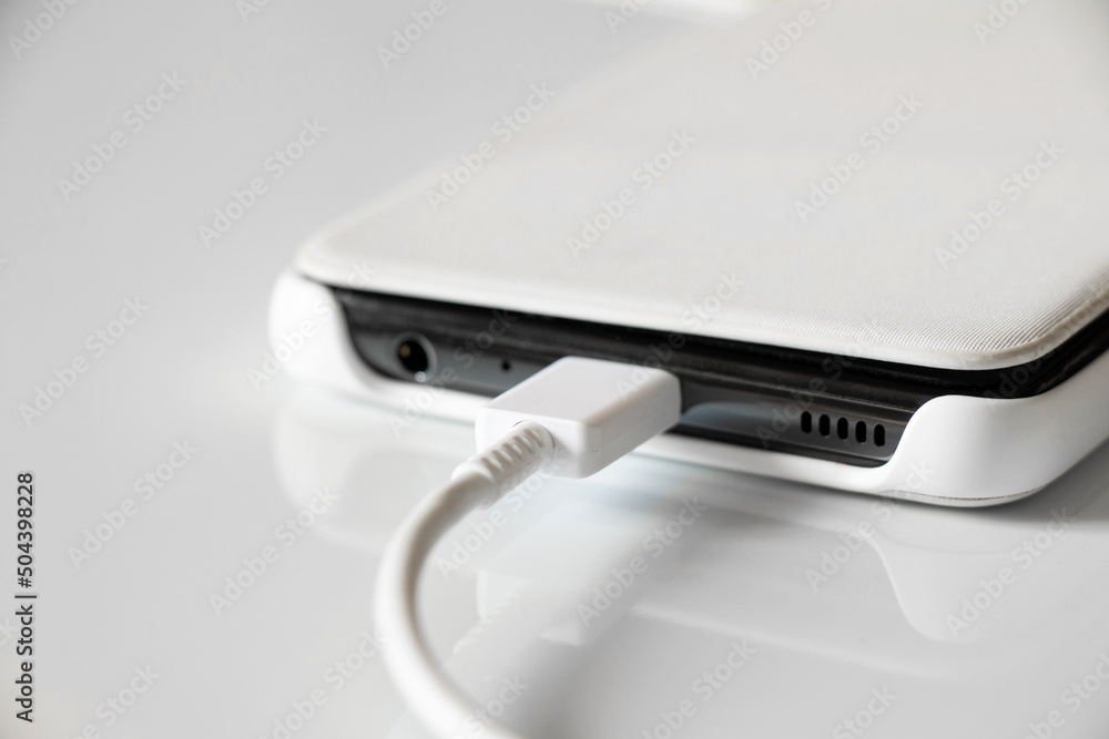White mobile phone in a case lies on recharging on a white background, USB cable and phone close up, fast phone charging White mobile phone in a case lies on recharging on a white background, USB cable and phone close up, fast phone charging