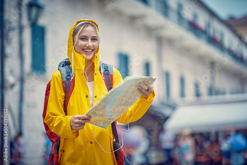 Billede på lærred Smiling  woman with a yellow raincoat and map on the street while enjoying a walk through the city on a rainy day