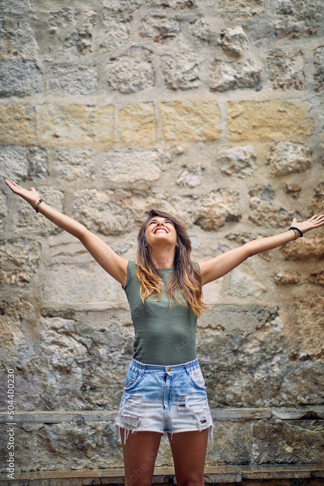 Young brunette woman with open arms standing and looking up in front of stone wall building. Happiness, freedom, contentment, summer concept. Young brunette woman with open arms standing and looking up in front of stone wall building. Happiness, freedom, contentment, summer concept.