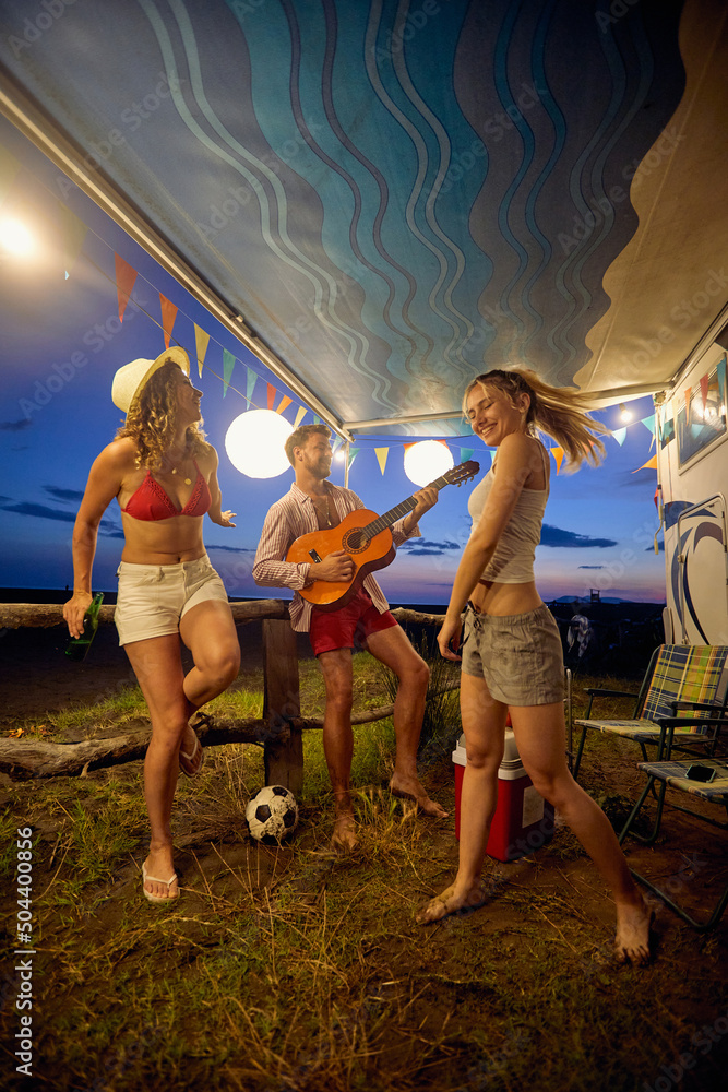 Friends celebrating in front of camper, man playing guitar women dancing, summertime camping. Fun, togetherness, travel concept.
