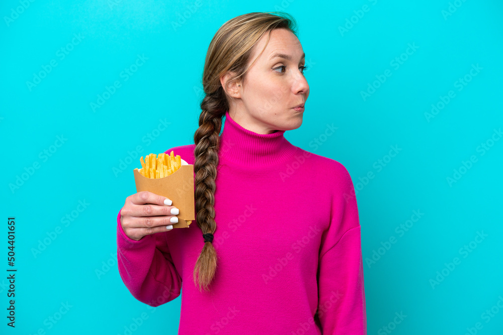 Young caucasian woman holding fried chips isolated on blue background looking to the side Young caucasian woman holding fried chips isolated on blue background looking to the side