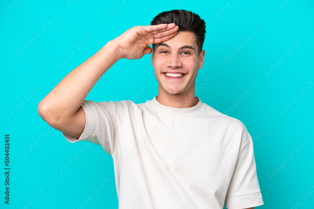 Young handsome caucasian man isolated on blue bakcground saluting with hand with happy expression Young handsome caucasian man isolated on blue bakcground saluting with hand with happy expression