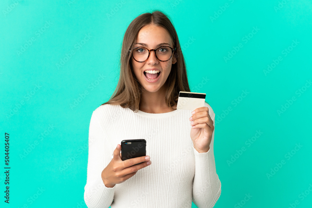 Young woman over isolated blue background buying with the mobile and holding a credit card with surprised expression Young woman over isolated blue background buying with the mobile and holding a credit card with surprised expression