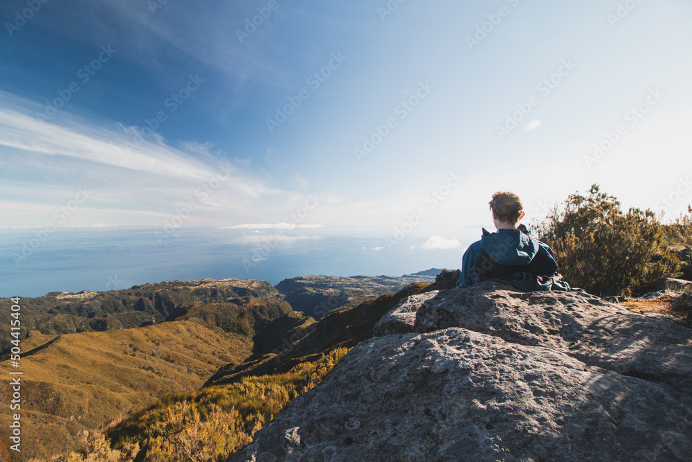 Blonde hiker takes a break on a rock overlooking the Atlantic Ocean and the Madeira Valley from the slopes of Pico Ruivo on the Portuguese island. Take a seat