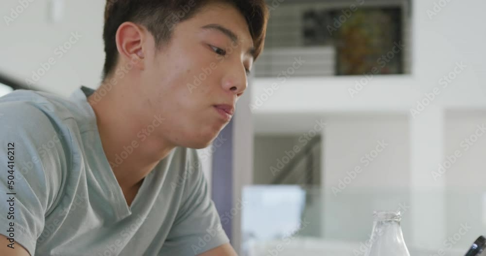 Asian male teenager sitting at table with smartphone and having breakfast Asian male teenager sitting at table with smartphone and having breakfast
