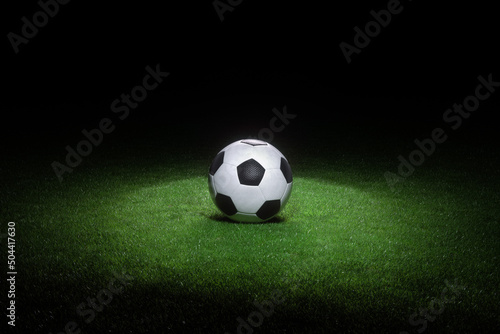 Black and white leather football ball laying on fresh green grass highlighted by spotlight.