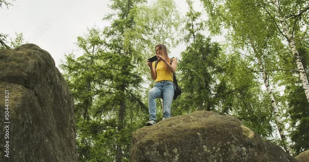 young girl climbed a hill and takes pictures in the forest on her smartphone, standing on a stone. Green trees on the background, bottom view young girl climbed a hill and takes pictures in the forest on her smartphone, standing on a stone. Green trees on the background, bottom view