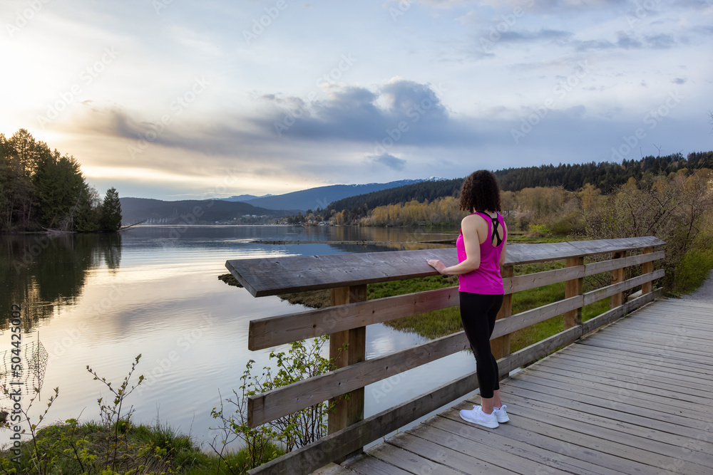 Woman Walking on a Wooden Path with green trees in Shoreline Trail, Port Moody, Greater Vancouver, British Columbia, Canada. Trail in a Modern City during a Cloudy Sunset.