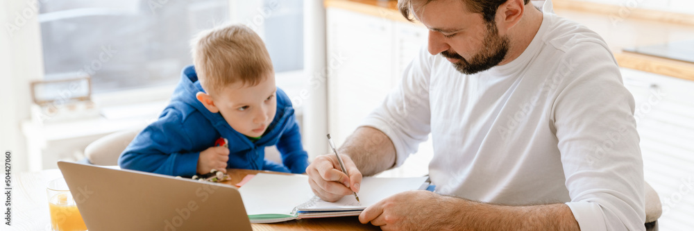 White father and son drawing and using laptop in kitchen at home White father and son drawing and using laptop in kitchen at home