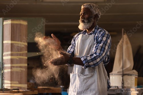 African american mature carpenter dusting sawdust on hands in workshop photo