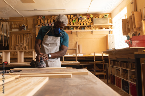 African american mature carpenter in facer mask using sander on plank at workbench in workshop