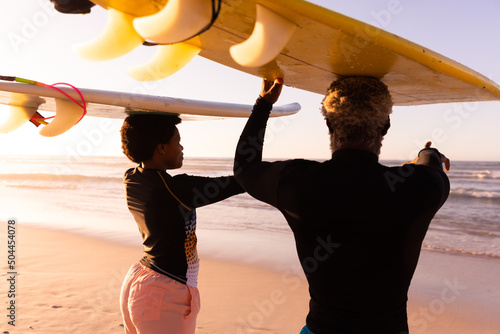 African american couple carrying surfboards on heads looking at sea against clear sky at sunset