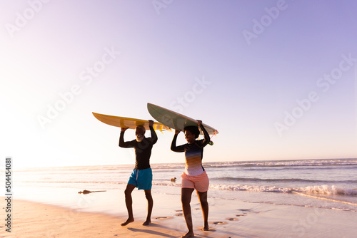 African american couple carrying surfboards on heads while walking against sea and clear sky