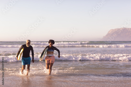 Carefree african american couple with surfboards running amidst waves against clear sky, copy space