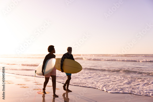 African american couple with surfboards walking at shore against clear sky at sunset , copy pace