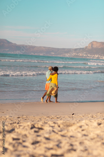 African american couple with arms around walking at shore against mountain and blue sky, copy space