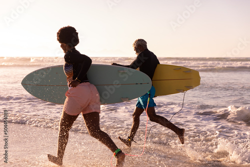 Cheerful african american couple with surfboards running in sea against clear sky at sunset