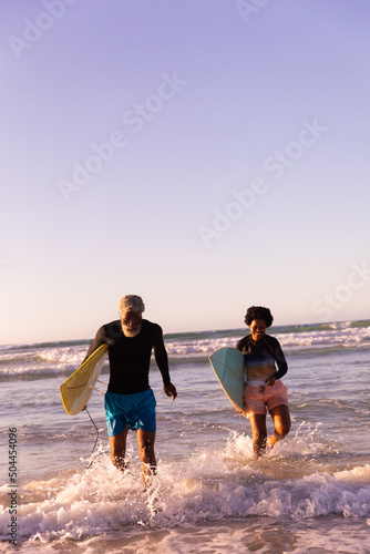 Happy african american couple with surfboards walking in sea against clear sky at sunset, copy space