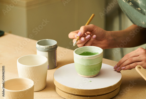 Close-up of girl painting clay mug with glaze. Woman coloring pottery in workshop with a paintbrush. Painter in green apron glazing clay pot.