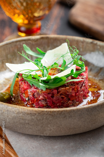 Beef tartare with dried tomatoes decorated with parmesan and arugula is served in a plate on a wooden table, close up
