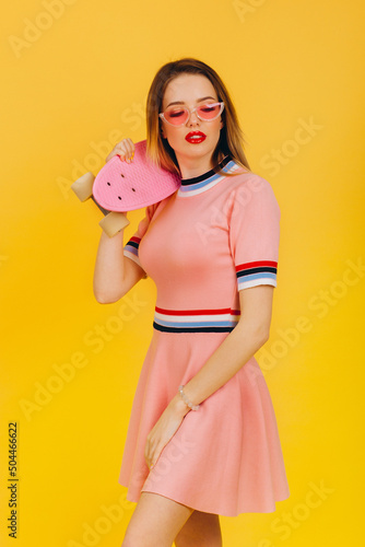 Portrait of happy smiling blonde girl hold pink skateboard on yellow background. People summer vacation rest lifestyle concept. Posing in pink short dress and sunglasses