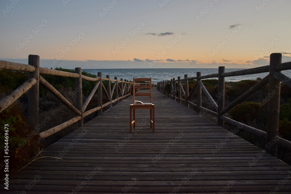 Bridge on the beach overlooking the sea, with a chair and a book whose pages flutter in the wind. Horizontal