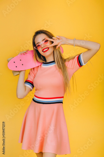 Urban lifestyle. Portrait of happy smiling blonde girl on yellow background. She holds a skateboard with one hand and shows the peace sign with the other. Happy lifestyle