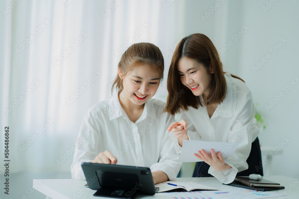 Two young Asian business women meet to analyze the financial chart at the office to discuss the financial situation at the company. A partner sits at a desk with modern documents and equipment. Two young Asian business women meet to analyze the financial chart at the office to discuss the financial situation at the company. A partner sits at a desk with modern documents and equipment.