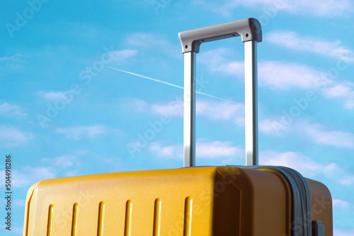 Yellow travel suitcase against a blue sky with an airplane trail photo