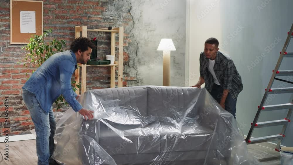 Black men cover new sofa with foil to protect from paint drops and sit down in room. Brothers prepare room for repairing. Home reconstruction organization Black men cover new sofa with foil to protect from paint drops and sit down in room. Brothers prepare room for repairing. Home reconstruction organization
