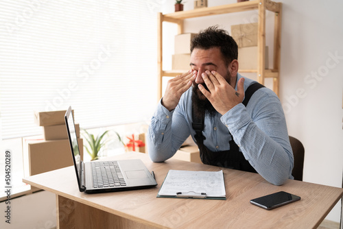tired indian male small business owner working at laptop, online store delivery worker has vision problems, businessman working at home office, sme