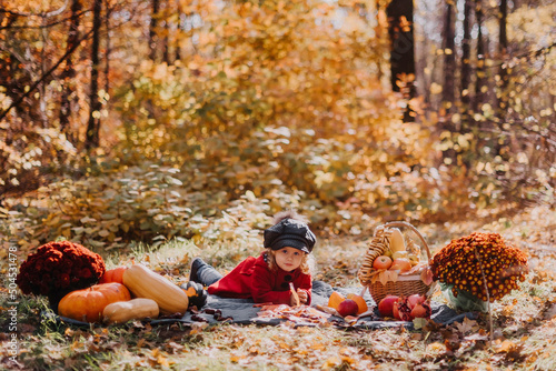Fototapeta Naklejka Na Ścianę i Meble -  Little girl in a red coat sits in an autumn forest withe a basket of autumn fruits