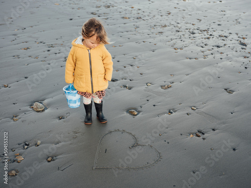 Young girl stands on a beach looking at a heart drawn in the sand.