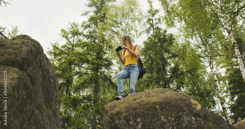 young girl climbed a hill and takes pictures in the forest on her smartphone, standing on a stone. Green trees on the background, bottom view