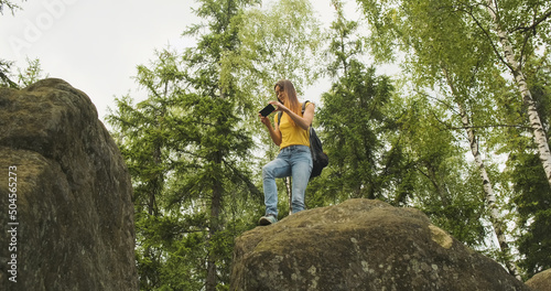 young girl climbed a hill and takes pictures in the forest on her smartphone  standing on a stone. Green trees on the background  bottom view