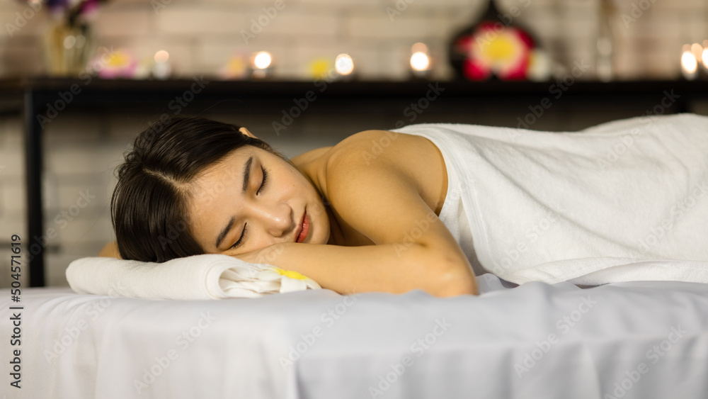 Closeup Asian young beautiful sexy happy female spa customer covered by white clean bath towel laying down resting relaxing closed eyes smiling on massaging bed putting bloom orchid flower on ear Closeup Asian young beautiful sexy happy female spa customer covered by white clean bath towel laying down resting relaxing closed eyes smiling on massaging bed putting bloom orchid flower on ear