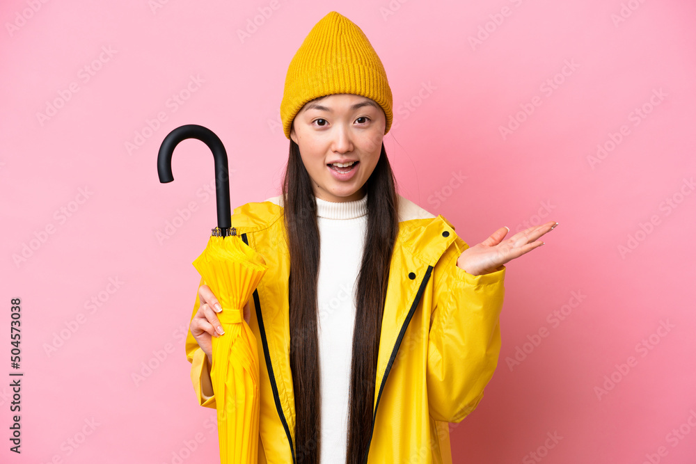 Young Chinese woman with rainproof coat and umbrella isolated on pink background with shocked facial expression Young Chinese woman with rainproof coat and umbrella isolated on pink background with shocked facial expression