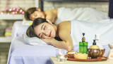 Closeup shot Asian young sexy female spa customer covered by white clean bath towel lay down on massaging bed with flowers smiling look at camera while girlfriend lying beside in blurred background