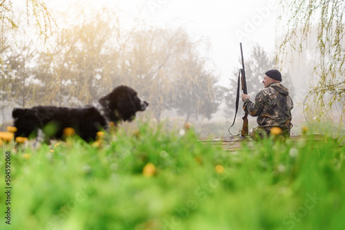 Male hunter sitting on a footbridge on a background of sunrise and forest with a lake in the foreground blurred dog spaniel