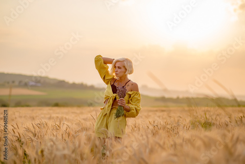Pretty young woman in yellow among rural field with golden oat field on sunset background