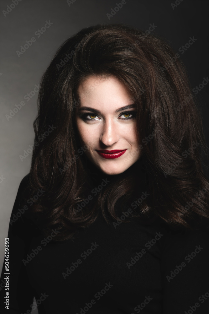 Beautiful brunette woman with long dark fluffy, wavy hair and green eyes studio portrait. Model with evening make-up and red lipstick wearing dark clothes in dark studio background looking to camera