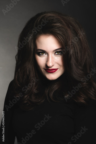 Beautiful brunette woman with long dark fluffy, wavy hair and green eyes studio portrait. Model with evening make-up and red lipstick wearing dark clothes in dark studio background looking to camera