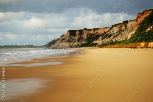 Cliffs on the shore, yellow sand beach and the sea water with calm waves. Horizon cloudy with light of early morning.