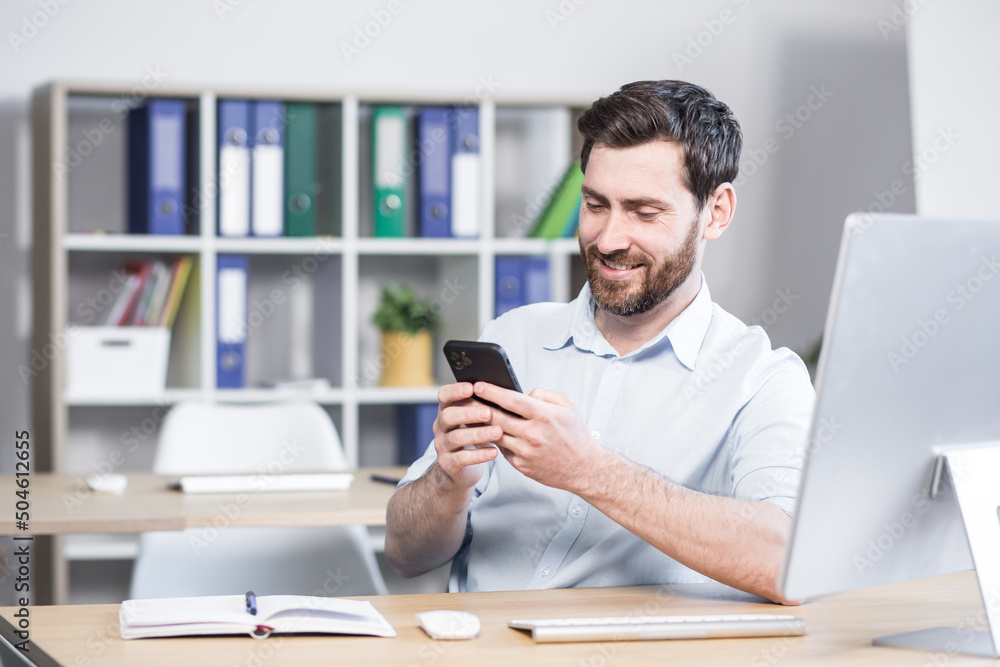 happy handsome office worker sitting at a computer desk in the office using a mobile phone. Cheerful bearded business man entrepreneur employee. browses smartphone reads messages chats communicates happy handsome office worker sitting at a computer desk in the office using a mobile phone. Cheerful bearded business man entrepreneur employee. browses smartphone reads messages chats communicates