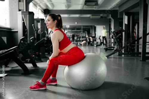A beautiful girl is sitting on a fitball. Girl in a red suit in the gym.